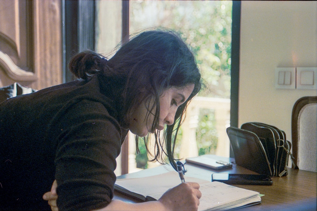 A student bent over a notebook at a desk by a window, writing late in the day.